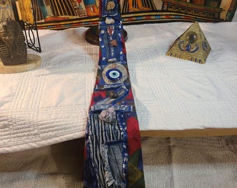 May include: A handmade necktie with a unique design. The tie features a patchwork of blue denim, red, and green fabrics, adorned with various embellishments including a blue eye charm and small stones. The text "JOHN HENRY" is visible on a small black label.