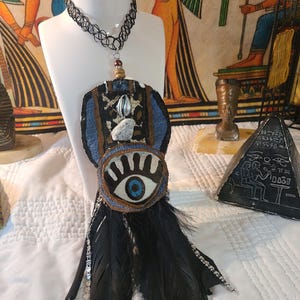May include: A unique necklace featuring a black choker, a pendant with an eye design, and black feathers. The pendant has a blue eye, a denim-like fabric, and a seashell accent. The necklace is displayed on a white mannequin.
