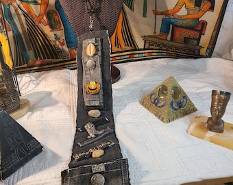 May include: A denim necklace with a black cord, adorned with decorative elements. The pendant features a pocket design with a yellow cowboy boot, stones, and metal buttons. The necklace is displayed on a mannequin.