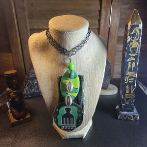 May include: A black choker necklace with a green and black pendant featuring a comb and a shell. The pendant is layered with green and black fabric and has a green bead at the top.