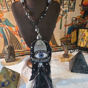 May include: A unique necklace featuring a black pendant with a blue eye design, adorned with silver-colored embellishments and black feathers. The necklace has a beaded chain with black and white beads. The pendant is displayed on a brown bust.
