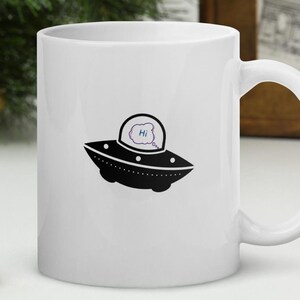 May include: White ceramic mug with a black and white illustration of a flying saucer with a speech bubble that says "Hi".