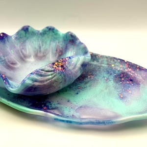 May include: A decorative serving set with a clam shell-shaped bowl and an oval platter. The set is swirled with blue, teal, and purple, with glitter accents. The bowl sits on the platter. Measurements are in centimetres.