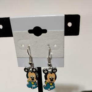 May include: A pair of silver dangle earrings featuring a cartoon image of Mickey Mouse wearing a blue shirt. The earrings have clear rhinestones on the ears and around the image.