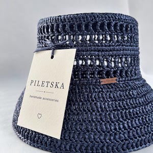 May include: A dark navy blue crocheted bucket hat with an open-weave pattern. A cream-colored rectangular paper tag is attached to the hat with a thin string, featuring the text PILETSKA handmade accessories and a small heart symbol.