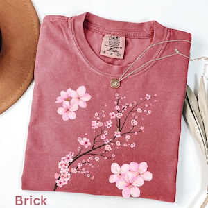 May include: A brick-colored t-shirt with a floral design. The shirt features a pink cherry blossom branch and flowers. A silver necklace with a pendant is draped across the shirt. The tag reads "Comfort Colors".