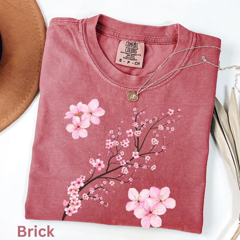 May include: A brick-colored t-shirt with a floral design. The shirt features a pink cherry blossom branch and flowers. A silver necklace with a pendant is draped across the shirt. The tag reads "Comfort Colors".