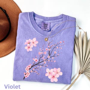 May include: A violet-colored Comfort Colors t-shirt featuring a floral design of pink cherry blossoms on a branch. The shirt has a crew neck and short sleeves. A gold necklace with a pendant rests on the shirt. The word "Violet" is printed at the bottom.