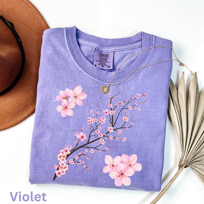 May include: A violet-colored Comfort Colors t-shirt featuring a floral design of pink cherry blossoms on a branch. The shirt has a crew neck and short sleeves. A gold necklace with a pendant rests on the shirt. The word "Violet" is printed at the bottom.
