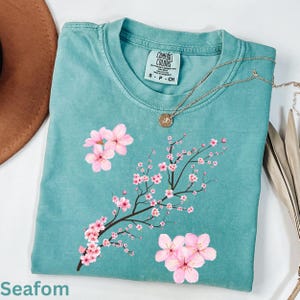 May include: Seafoam green t-shirt with a floral design. The shirt features a branch with pink cherry blossoms and the word "Comfort Colors" on the tag. A gold necklace with a pendant is draped across the shirt.