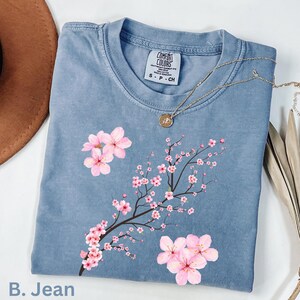 May include: A blue Comfort Colors t-shirt featuring a floral design of pink cherry blossoms on a branch. The shirt has a crew neck and short sleeves. A gold necklace with a pendant is draped on the shirt. The text "B. Jean" is at the bottom.