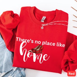 May include: A red sweatshirt with the text "There's no place like home" and an illustration of red glitter heels. A silver necklace with a sunburst pendant is on the sweatshirt. The word "Red" is in a red box in the bottom right corner.