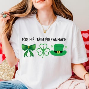 May include: White t-shirt with the text "PÓG MÉ, TÁIM ÉIREANNAC". The shirt features green graphics of a bow, a shamrock, and a leprechaun hat. The design is perfect for St. Patrick's Day celebrations.