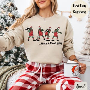 Może przedstawiać: Beżowa bluza z nadrukiem czterech zielonych postaci w czerwonych ogrodniczkach i czapkach Świętego Mikołaja, z napisem "...that's it I'm not going." Osoba ma na sobie czerwono-białą piżamę w kratę i trzyma kubek z motywem Świętego Mikołaja.