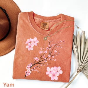 May include: A yam-colored Comfort Colors sweatshirt featuring a floral design of pink cherry blossoms on a branch. A gold necklace with a pendant is draped across the shirt. The shirt is folded on a white surface.