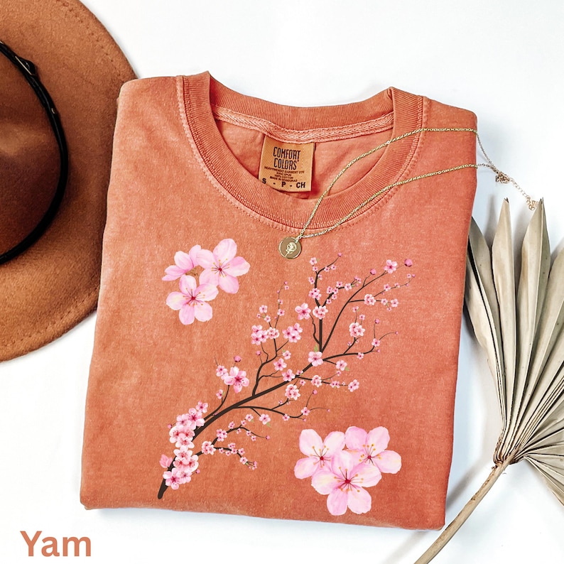 May include: A yam-colored Comfort Colors sweatshirt featuring a floral design of pink cherry blossoms on a branch. A gold necklace with a pendant is draped across the shirt. The shirt is folded on a white surface.
