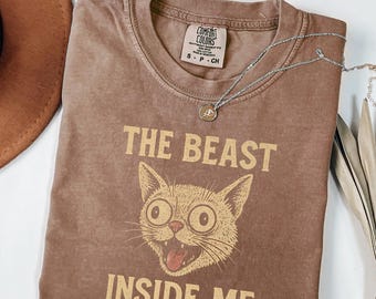 May include: Espresso-colored t-shirt with the text "THE BEAST INSIDE ME" and a cartoon cat graphic. The shirt has a crew neck and short sleeves. A silver necklace with a pendant is draped across the shirt.