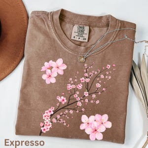 May include: A brown Comfort Colors t-shirt with a floral design of pink cherry blossoms on a branch. The word "Expresso" is printed at the bottom. A silver necklace with a pendant is draped across the shirt.