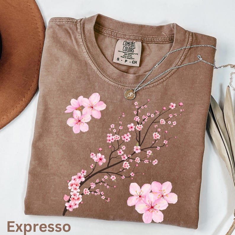 May include: A brown Comfort Colors t-shirt with a floral design of pink cherry blossoms on a branch. The word "Expresso" is printed at the bottom. A silver necklace with a pendant is draped across the shirt.