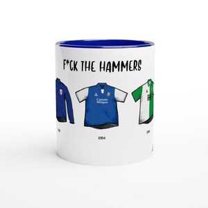 Peut inclure: Mug en céramique blanche avec un intérieur bleu et le texte "F*ck the Hammers". La tasse présente des illustrations de trois maillots de football de différentes années. Les maillots sont bleus, blancs et verts.