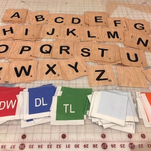Könnte beinhalten: Ein Satz Holzbuchstaben mit schwarzer Beschriftung, einschließlich des Alphabets und eines Herzens. Die Kacheln sind in Reihen auf einer gerasterten Oberfläche angeordnet. Unter den Kacheln befinden sich Stapel bunter Stoffquadrate mit den Buchstaben DW, DL, TL und TW.