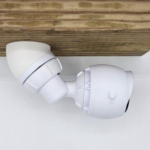 以下が含まれることがあります： 木製の表面に取り付けられた旋回式マウントを備えた白いセキュリティカメラ。カメラは前面にレンズがあり、側面にU字型のロゴがあります。