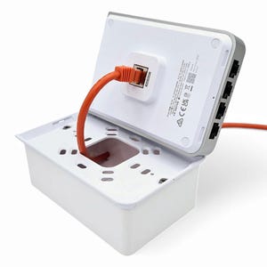 Puede incluir: Una placa de pared blanca con un cable Ethernet naranja conectado a un puerto. La placa está abierta y el cable pasa por la abertura. La placa tiene una serie de pequeños agujeros alrededor del borde.