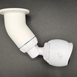 Puede incluir: Cámara de seguridad blanca con un soporte de montaje blanco. La cámara está montada en el soporte y apunta hacia la derecha. La cámara tiene una lente negra y un logotipo U blanco en la parte delantera.