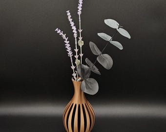 May include: A bronze-colored, open-slatted vase holds lavender-colored sprigs and gray eucalyptus leaves. The vase has a rounded shape with vertical cutouts, creating a decorative and modern design. The arrangement is set against a dark background.
