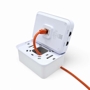 Op de afbeelding: Een witte plastic doos met een afneembaar deksel. Het deksel is open en een oranje ethernetkabel is aangesloten op een poort aan de achterkant van de doos. De doos heeft twee andere poorten aan de achterkant.