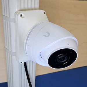 May include: A white security camera with a black lens mounted on a white wall. The camera has a U-shaped logo on the side.
