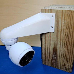 May include: A white security camera mounted on a wooden post with a white bracket. The camera has a round lens and a white casing.