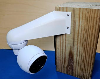 May include: A white security camera mounted on a wooden post with a white bracket. The camera has a round lens and a white casing.