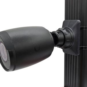 Puede incluir: Una cámara de seguridad negra con una cubierta protectora está montada en un poste negro. La cámara es cilíndrica con una lente visible en la parte delantera. Está sujeta al poste con un soporte negro y dos correas negras.