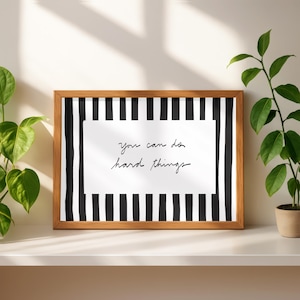 May include: A framed print with a wooden frame. The print features a white rectangle with the handwritten text "you can do hard things" against a black and white striped background. The frame is brown.
