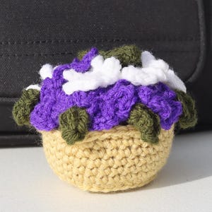Peut inclure: Un panier de fleurs crocheté avec des fleurs violettes, blanches et vertes. Le panier beige est placé devant un sac noir. Les fleurs sont disposées de manière dense, créant un motif texturé. Un article fait main charmant.