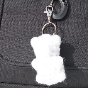 May include: A black backpack with a white, crocheted teddy bear keychain attached. The keychain is hanging from a black carabiner clip. The backpack has a zippered pocket and red accents.