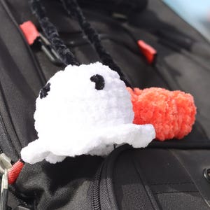 Op de afbeelding: Een witte gehaakte spookvormige bedel met zwarte ogen en een rode pompom-accent, bevestigd aan een zwarte rugzak. De geest heeft een gerimpelde rand. De rugzak is zwart met rode ritstrekkers en zilveren hardware.