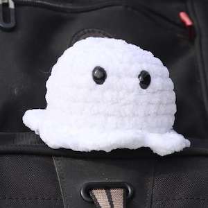 May include: A white crocheted ghost with black eyes is attached to a black rucksack. The ghost has a rounded body and a wavy bottom edge. The rucksack has multiple compartments, red zip pulls, and a handle at the top.