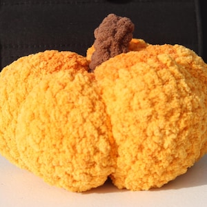 May include: A plush, handmade pumpkin in shades of orange and yellow, with a textured, fuzzy appearance. The pumpkin has a brown stem and is likely a decorative item for fall or Halloween.