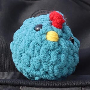 May include: A handmade, teal blue crocheted chick with black button eyes, a yellow beak, and a red comb. The plush toy is round and fluffy, ideal for a child's toy collection.