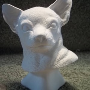 May include: A white ceramic bust of a dog with large, upright ears and detailed fur texture. The sculpture features a smooth, matte finish and captures the head and neck of the animal in a forward-facing position with alert eyes.
