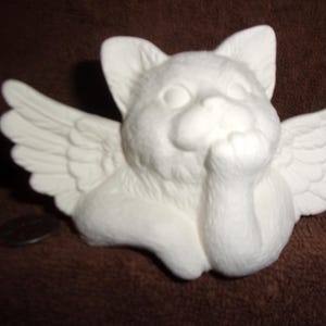 May include: A white ceramic figurine of a cat with feathered wings extending from its sides. The cat is depicted in a resting position with one paw raised toward its chin, featuring detailed fur texture and smooth, unpainted matte white surfaces.