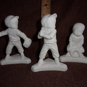 May include: Three unpainted ceramic baseball figurines. One figure is throwing a baseball, another is holding a bat, and the third is kneeling with a baseball cap. All are wearing baseball uniforms and caps.