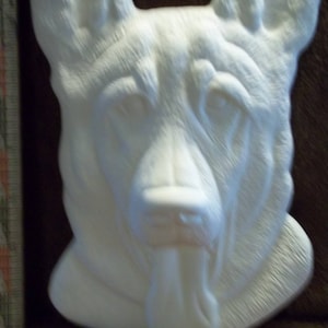 May include: A white, unpainted ceramic German Shepherd dog head wall plaque with textured fur details and a protruding tongue. A ruler is visible on the left side, showing the piece measures approximately 8 inches in total height.
