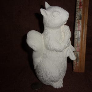 May include: A white ceramic squirrel figurine holding an acorn, positioned next to a wooden ruler showing a height of approximately 7 inches. The squirrel has textured fur details, pointed ears, and a bushy tail, standing upright on a flat surface.
