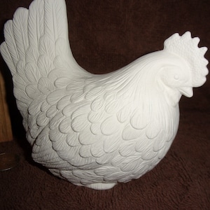 May include: A white ceramic chicken figurine with detailed feather textures, a prominent comb, and a wattle. A wooden ruler is positioned vertically next to the figurine, showing a height measurement of approximately 6 inches for the chicken.