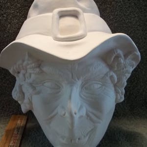May include: A white ceramic bisque witch head sculpture wearing a pointed hat with a buckle. The sculpture features detailed facial features including a pointed nose and textured hair. A wooden ruler is visible, showing a scale of 6 inches.