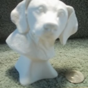 May include: A white ceramic dog bust sculpture featuring floppy ears and a smooth finish. The figurine is positioned next to a silver quarter coin and a wooden ruler, which shows the base of the sculpture is approximately 2.5 inches wide.