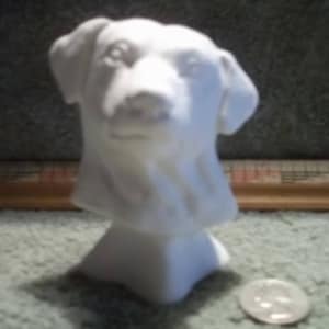 May include: A white ceramic dog head sculpture. The sculpture is a bust of a dog's head, with detailed features. The dog's head is mounted on a pedestal base. The sculpture is displayed on a green surface.